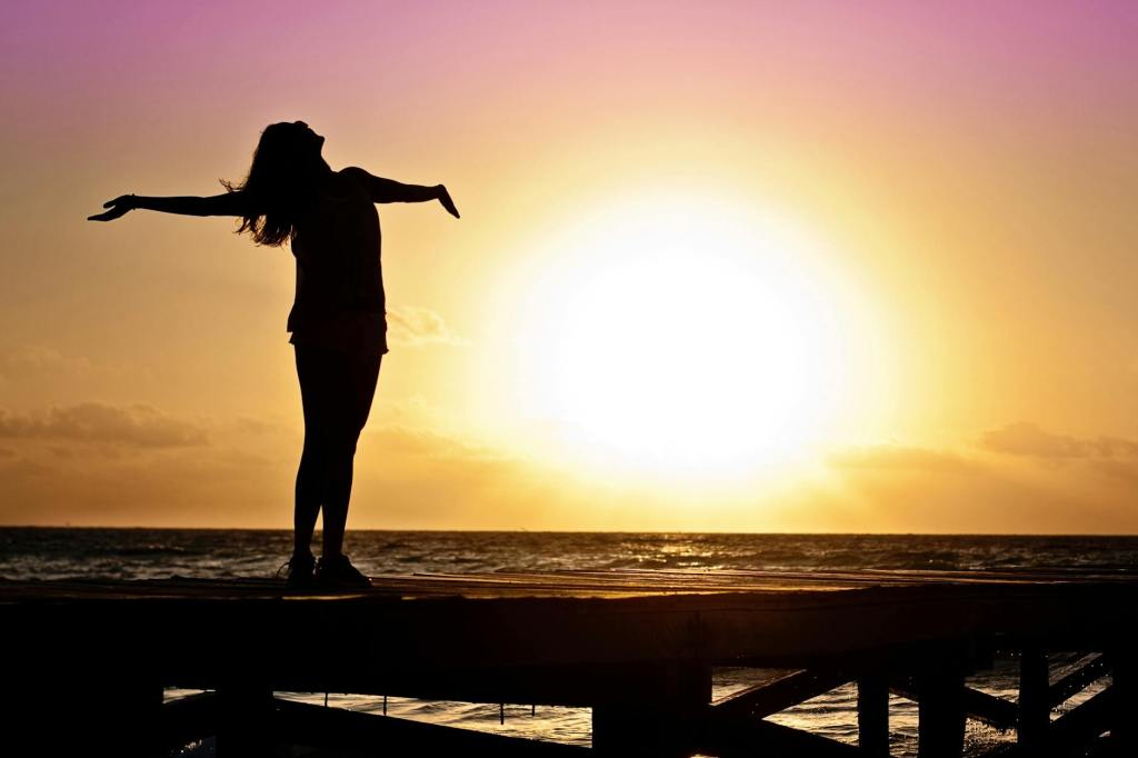 A woman standing on a doc in the sunset with her arms spread open